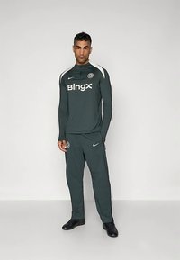 Zelená tepláková súprava s čiernymi akcentmi, ktorú zdobí vysoký golier, logo na hrudi a elastické manžety a pás. Športová obuv dotvára celý vzhľad.