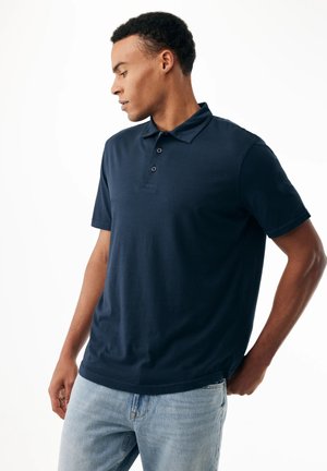 Joven con una camisa polo de manga corta azul marino y jeans azul claro, mirando hacia abajo a su izquierda con las manos cerca de los bolsillos.