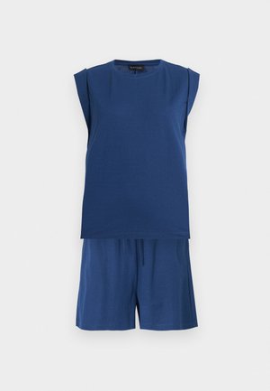 Kobieta ubrana w granatowy bezrękawnik i szorty, białe sportowe buty oraz czarne okulary przeciwsłoneczne, stoi z jedną ręką w kieszeni na białym tle.