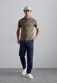 Olive green polo shirt, navy trousers, white sneakers, and a light blue cap. Model stands against a plain gray background.