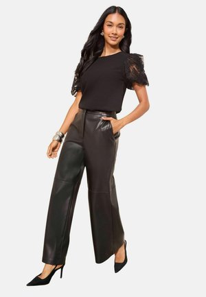 Svart topp med puffärmar i spets, kombinerad med högmidjade, vida svarta läderbyxor. Modellen bär svarta spetsiga klackskor.