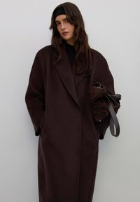 Bruine overjas met een brede kraag, zachte textuur. Model houdt een donkerbruine tas met leren bandjes vast. Simpel, minimalistisch design.