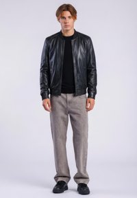 Junger Mann in schwarzer Lederjacke, schwarzem Hemd, weiten grauen Hosen und schwarzen Schuhen, der vor einem schlichten hellen Hintergrund steht.