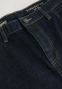 Jeans de mezclilla azul oscuro con un corte recto, que presentan costuras amarillas en contraste, un cierre con botón y una etiqueta que dice "Supply 34."