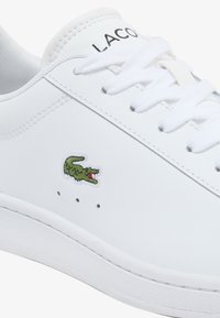 Sneaker in pelle bianca con punta arrotondata, lacci bianchi e logo Lacoste. Presenta un coccodrillo ricamato verde e piccoli fori di ventilazione vicino alla suola.