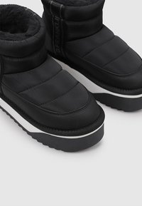 Botas de invierno acolchadas negras con un forro interior de felpa, suela blanca y detalles texturizados; presentan un logo en el lateral.
