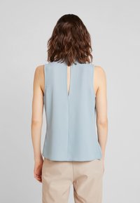 Top senza maniche di colore azzurro chiaro con schiena a chiave, realizzato in tessuto morbido, caratterizzato da una vestibilità comoda e un orlo arrotondato. Abbinato a pantaloni beige.
