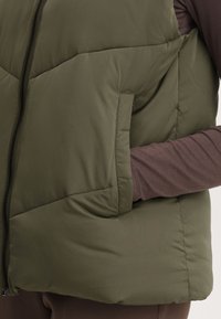 Gilet en duvet vert olive avec fermeture éclair, présentant un motif matelassé en losanges et des poches latérales. Porté par-dessus un haut à manches longues marron.