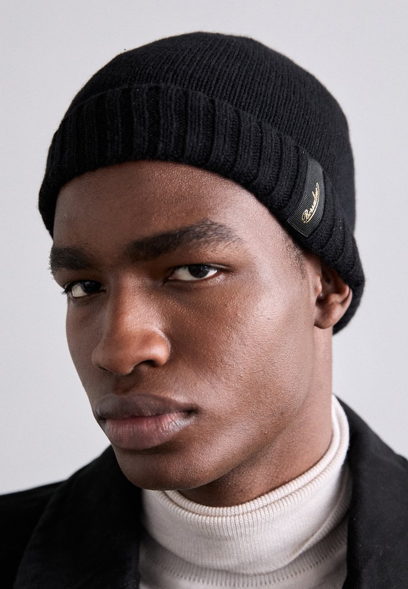 Gorro de malha preto com textura canelada e um patch de logótipo. Usado com uma camisola de pescoço alto clara e um casaco escuro, apresenta um close-up do rosto.