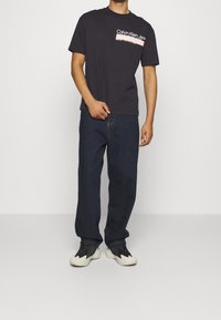Hombre con camiseta negra de Calvin Klein Jeans, jeans de color azul oscuro y zapatillas en blanco y negro, de pie frente a un fondo claro y liso.