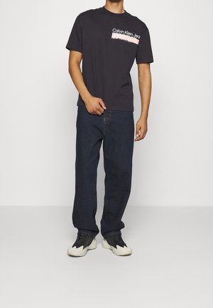 Hombre con camiseta negra de Calvin Klein Jeans, jeans de color azul oscuro y zapatillas en blanco y negro, de pie frente a un fondo claro y liso.