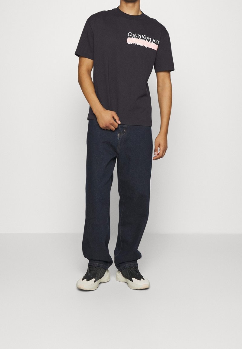 Hombre con camiseta negra de Calvin Klein Jeans, jeans de color azul oscuro y zapatillas en blanco y negro, de pie frente a un fondo claro y liso.
