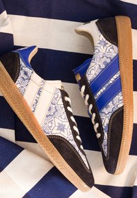 Paire de baskets noires, blanches et bleues avec motifs floraux et semelles en gomme sur un fond en tissu rayé bleu marine et blanc.