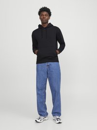 Black hooded sweatshirt with a kangaroo pocket, paired with loose-fitting blue jeans and multi-colored sneakers, set against a plain white background.