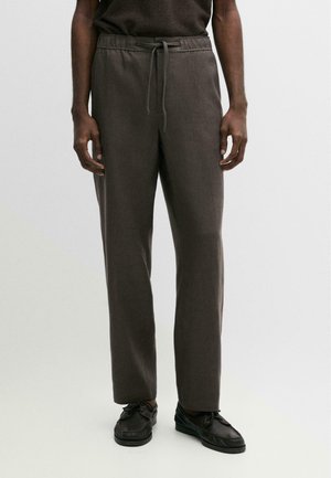 Uomo che indossa pantaloni marrone scuro con coulisse e scarpe in pelle nera, in piedi davanti a uno sfondo chiaro e uniforme.