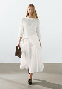Maglione bianco a maglia con maniche a tre quarti, sovrapposto a una gonna bianca fluida. Una borsa a tracolla in tessuto scuro e sandali platform neri completano il look.