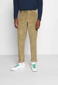 Tan kordbyxar med klassisk passform, rakt ben och uppvikta fållar, kombinerade med vita sneakers och gröna snörningar.