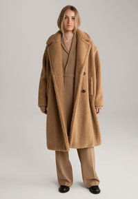 Langer, oversized brauner Mantel aus strukturiertem, plüschigem Stoff. Er hat große Revers, zwei Knöpfe und Vorderaschen. Gestylt über einem beigen Outfit.