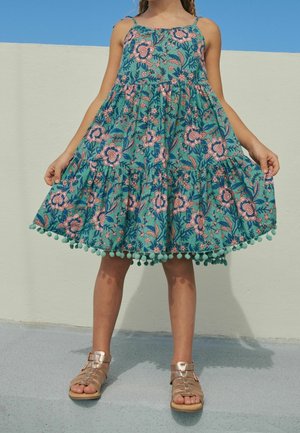 Dziewczyna w miętowej sukience w kwiaty z pomponikami na brzegu, trzymająca dół sukienki, stojąca na zewnątrz na tle beżowej ściany i błękitnego nieba.