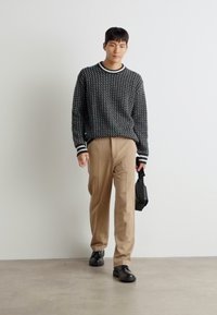 Sorte mønstrete genser med hvite detaljer, beige bukser og svarte sko; personen holder en liten svart veske mens de går.