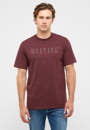 Junger Mann trägt ein kastanienbraunes Mustang-T-Shirt und blaue Jeans und steht vor einem schlichten hellen Hintergrund.
