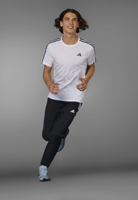 Weißes Adidas-T-Shirt mit schwarzen Streifen, kombiniert mit schwarzen Adidas-Hosen mit weißen Akzenten. Blaue Sportschuhe in Bewegung vor einem grauen Hintergrund.