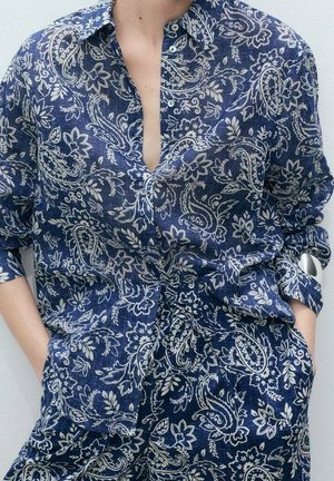 Chemise manches longues à motif paisley bleu et blanc et pantalon assorti, partiellement déboutonnée, portée avec les mains dans les poches.