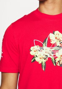Červená košile s krátkým rukávem, na které je velké bílé logo obrysově přeškrtnuté žlutými květinami a zelenými listy, nošena osobou.