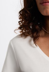 Witte V-hals top met een zachte, gladde textuur. Heeft korte mouwen en een subtiele vorm. Krullend donker haar gedeeltelijk zichtbaar. Zilver sieraad als accent.