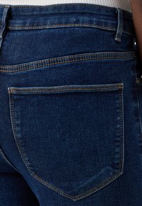 Σκούρο μπλε τζιν παντελόνι με εφαρμοστή σιλουέτα, με πέντε τσέπες και αντιθετική χρυσή ραφή κατά μήκος των ραφών και των τσεπών.