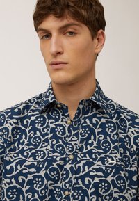 Blauwe katoenen shirt met een wit bloemenpatroon, met knoopsluiting en een klassieke kraag. Opmerkelijke details zijn een klein logo boven de zak.