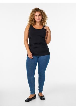 Lockenhaarige Frau steht auf weißem Hintergrund, trägt ein schwarzes ärmelloses Oberteil, blaue enge Jeans und schwarze Ballerinas.