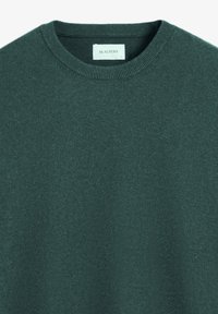 Suéter de cuello redondo en verde oscuro, hecho de un tejido suave. Presenta un logotipo sutil en el pecho y puños y cuello acanalados para un detalle adicional.