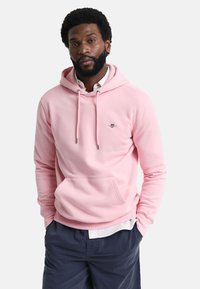 Felpa rosa con tasca frontale, cappuccio con coulisse e un piccolo logo sul petto. Il tessuto sembra morbido e testurizzato. Indossata sopra una camicia bianca.