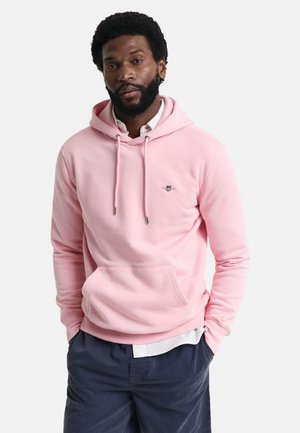 Sudadera rosa con bolsillo frontal, capucha con cordón y un pequeño logo en el pecho. La tela parece suave y texturizada. Lleva puesta sobre una camiseta blanca.