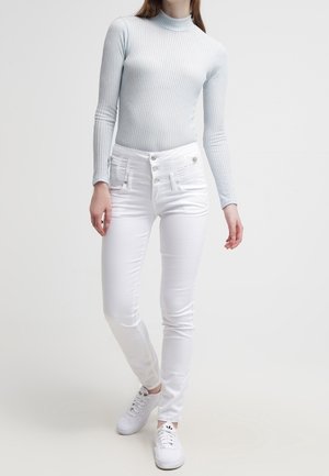 Person iført en lyseblå ribstrikket rullekrave, højtaljede hvide skinny jeans med flere knapper og hvide sneakers, stående med det ene ben krydset.