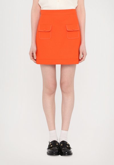 Mini-jupe orange vif avec deux poches plaquées à l'avant et des coutures blanches contrastées. Conçue avec une silhouette ajustée et une texture lisse.
