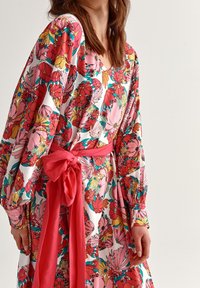 Vestido estampado floral en vibrante rosa, naranja y verde con mangas largas abullonadas y un cinturón rosa en la cintura, exhibiendo un diseño de escote en V.