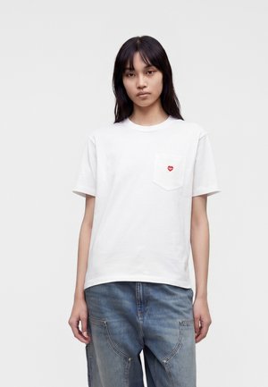 Ung kvinde med glat mørkt hår har en hvid t-shirt på med et lille rødt hjerte-logo på brystlommen og løse blå jeans.