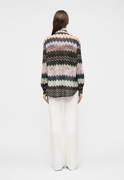 Γυναίκα με μακριά καστανά μαλλιά που φοράει πολύχρωμη μπλούζα μακρυμάνικη με ζιγκ-ζαγκ σχέδιο και άσπρο παντελόνι φαρδιάς γραμμής με διακριτικό σχέδιο σε σχήμα τσεβρόν, όρθια.