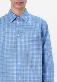 Blau karierte Hemd aus leichtem Stoff, mit weißen Knöpfen und einer einzelnen Brusttasche. Das Karomuster enthält schwarze und graue Linien.