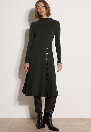 Mujer con vestido de punto verde oscuro de manga larga con botones delanteros y botas negras de tacón hasta la rodilla, de pie frente a una pared beige lisa.