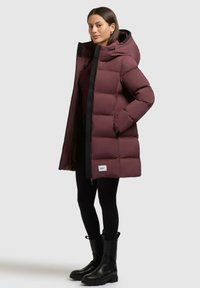 Giacca puffer bordeaux con cappuccio, dotata di zip frontale, tasche laterali e una trama compatta. Abbinata a leggings neri e stivali al ginocchio.
