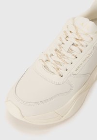 White sneaker with a smooth leather upper, textured accents, gold-flecked laces, and embossed branding on the side. Curved sole design.