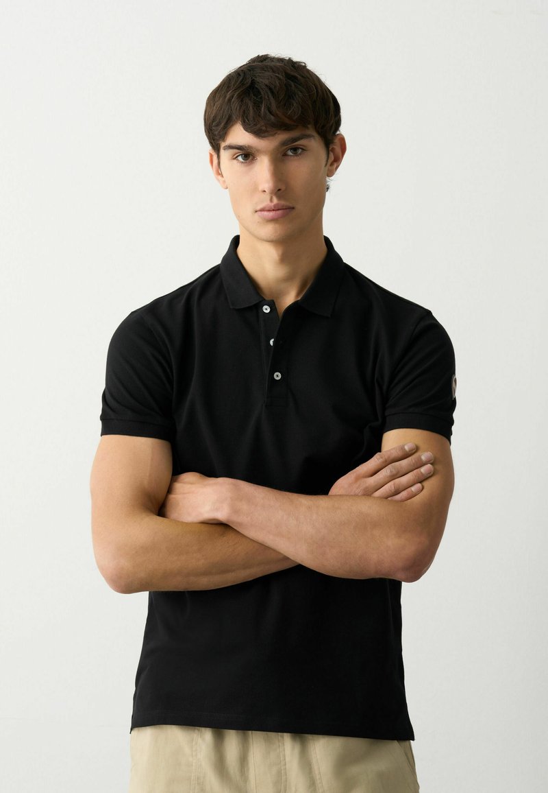 Camiseta polo ajustada negra con una textura suave, tapeta de tres botones y detalle del logo en la manga. El modelo tiene los brazos cruzados frente a un fondo liso.