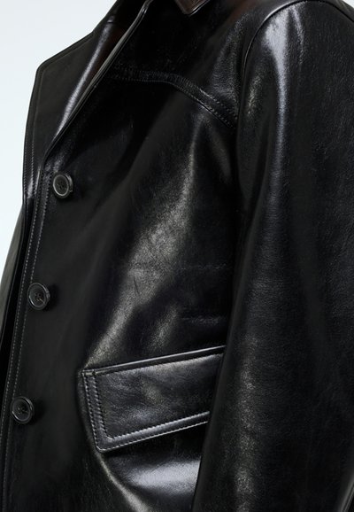 Black leather jacket with a shiny finish, featuring four buttons, a chest pocket, and structured lapels. Smooth texture and tailored fit.