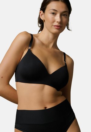 Kvinde iført sort trådløs bh og matchende højtaljede trusser med gyldne hoop-øreringe mod en ensfarvet lys baggrund.