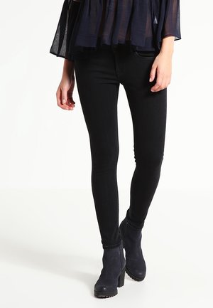 Person iført sorte skinny jeans, navy blå gennemsigtig top og mørkeblå hælede ankelstøvler står på hvid baggrund.