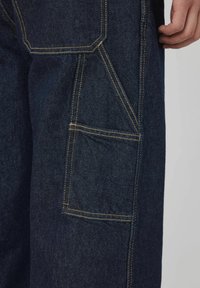 Pantalons cargo en denim bleu fonc é avec des coutures jaunes contrastantes. Comprend plusieurs poches, y compris une grande poche latérale avec un rabat.