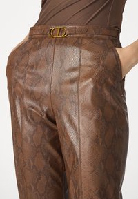 Bruine leren broek met een slangenhuidpatroon, voorzien van een riem met een gouden metalen gesp. De gladde textuur en zijzakken zijn zichtbaar.